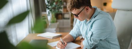 Photo of young student sitting on an office chair and writing at a desk in front of a window in a bright room