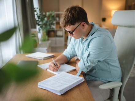 Photo of young student sitting on an office chair and writing at a desk in front of a window in a bright room