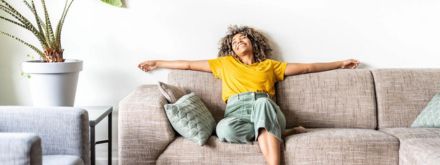 A woman in a yellow shirt and green pants relaxes with arms outstretched on a beige sofa in a bright, cozy living room. Decorative pillows and a large potted plant add comfort and style to the space.