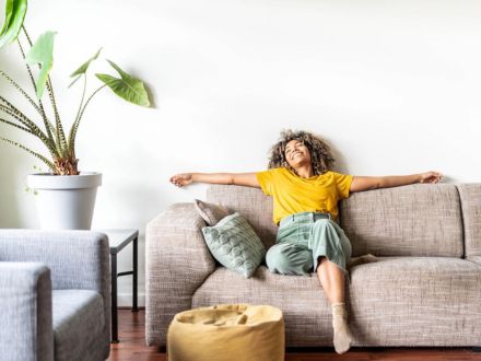 A woman in a yellow shirt and green pants relaxes with arms outstretched on a beige sofa in a bright, cozy living room. Decorative pillows and a large potted plant add comfort and style to the space.