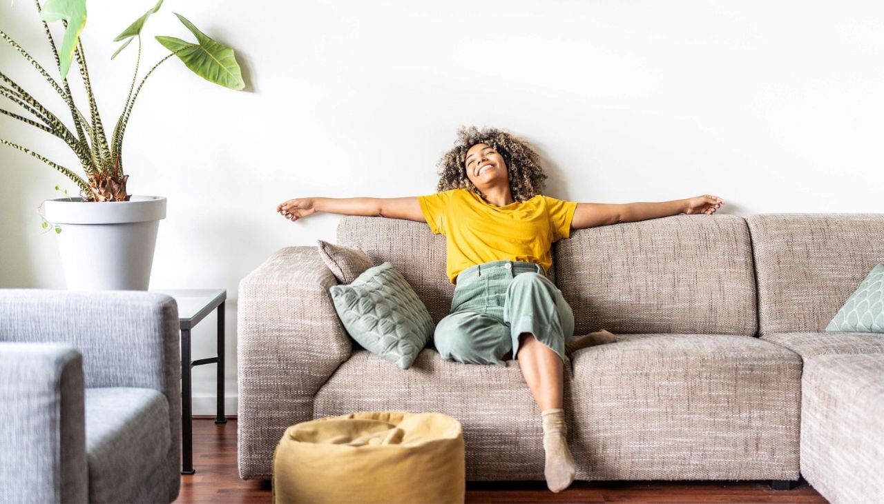 A woman in a yellow shirt and green pants relaxes with arms outstretched on a beige sofa in a bright, cozy living room. Decorative pillows and a large potted plant add comfort and style to the space.