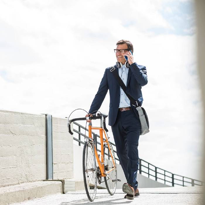 Man dressed in a business suit walking outdoors, holding a bicycle and talking on a smartphone. He has headphones around his neck and a shoulder bag.