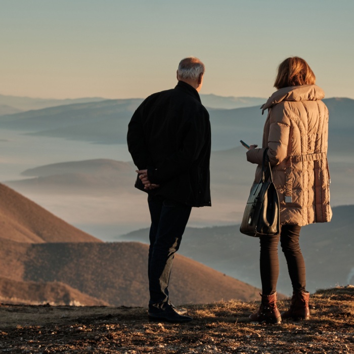 Two people in winter clothing overlook a misty mountain landscape at sunrise or sunset.